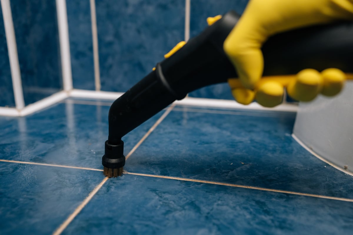 Close-up of person in yellow protective gloves operating powerful steam cleaner with brush attachment, deep cleaning blue ceramic bathroom tiles and grout lines for enhanced hygiene and sanitation.