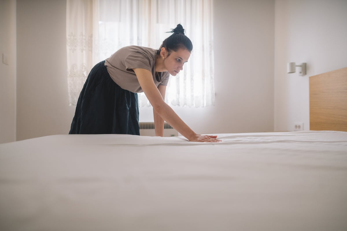 Female housekeeper tidying bed in hotel or apartment accommodation.