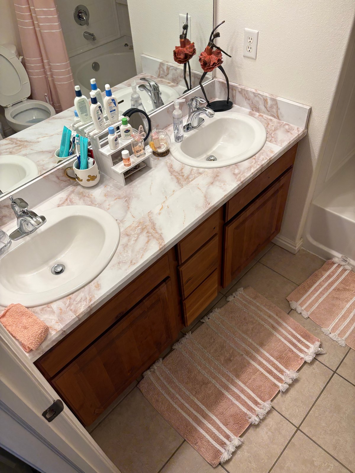 Modern bathroom with double sink vanity featuring pink marble countertop, various toiletries, and decorative coral flowers in vase