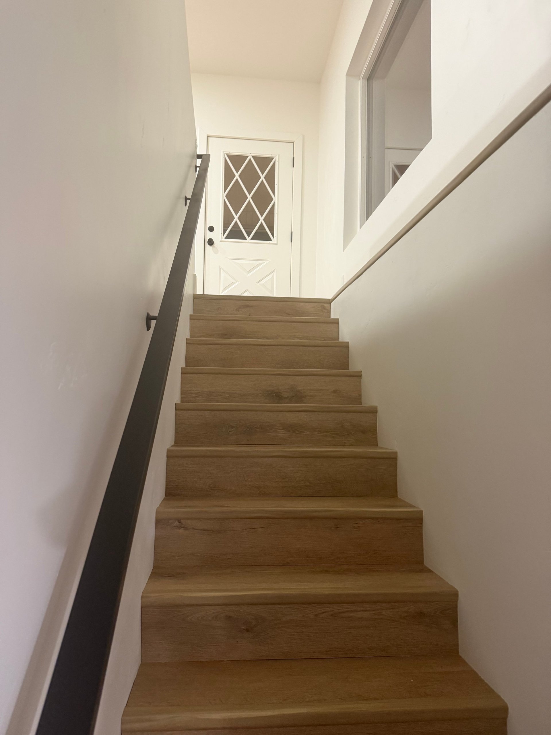 Wooden staircase with white walls and black handrail leading to white door with lattice window