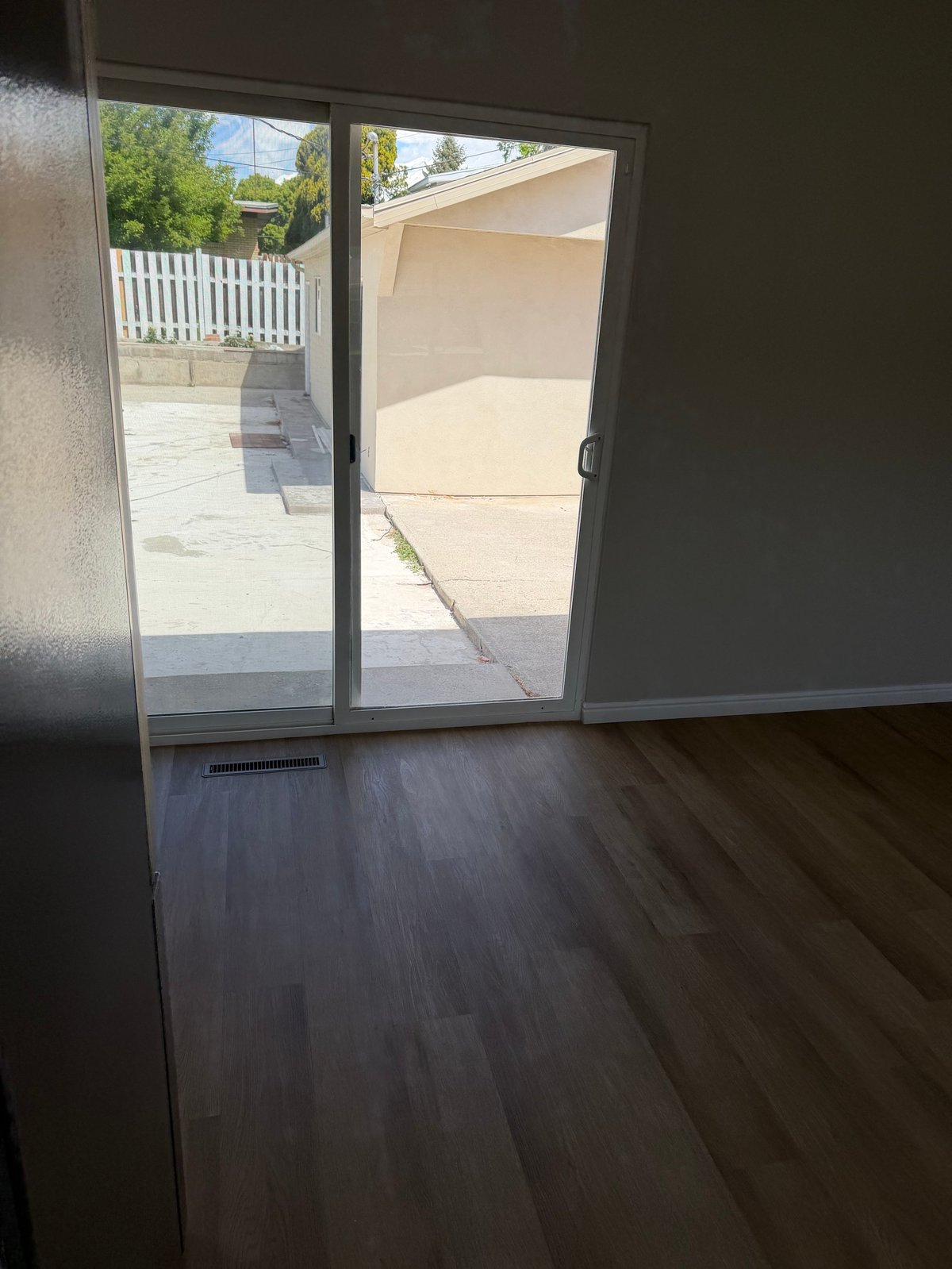 Empty room with dark hardwood floors and glass sliding patio doors opening to a sunny outdoor deck and driveway