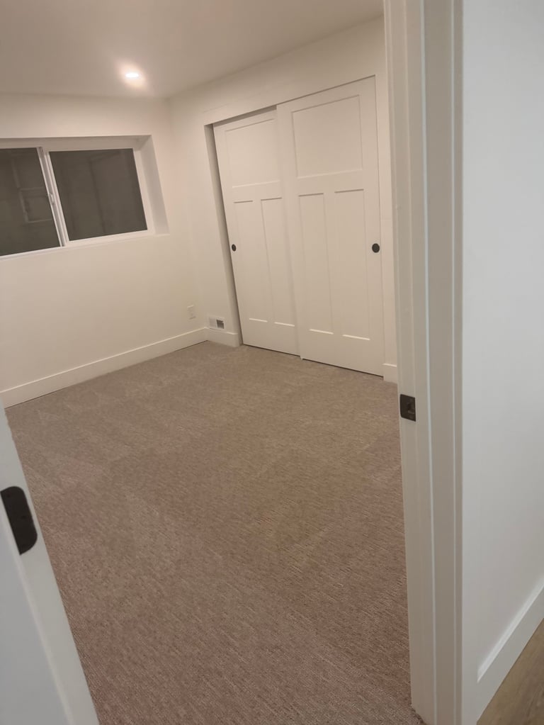 Empty bedroom with beige walls, white sliding closet doors, small window, and tan carpeted floor
